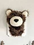 A plush bear head mounted on a white wooden plaque in a forest themed nursery. This stuffed bear wall mount is handmade by ClaraLoo with soft brown faux fur and crocheted accents.