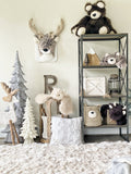 A plush deer wall mount in a woodland themed nursery. The stuffed deer head is made with soft brown and white fabric with crocheted ears. Other forest decor items include three tree sculptures and a brown bear stuffed animal.